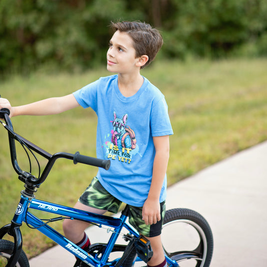 Child in a blue shirt with a graphic Frenchie design, standing next to a blue bicycle on a path with greenery in the background. The shirt says "is it time for PE yet?
