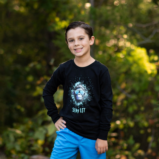 Child wearing a black long sleeve tee with a monkey graphic design and Stay Lit text, standing outdoors with greenery in the background.
