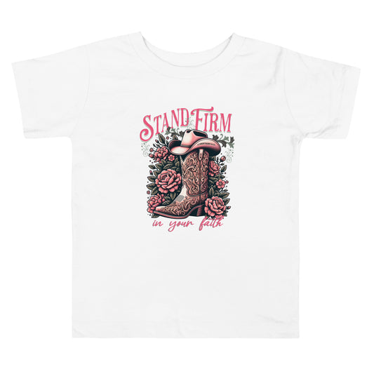 Christian graphic tee featuring a cowboy boot and floral design with the inspirational phrase “Stand Firm in Your Faith” on a pink background. Includes Bible verse 1 Corinthians 16:13 inscribed on the hat. Faith-based western apparel on white t-shirt.
