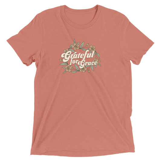 Colorful women's t-shirt laid flat, featuring delicate flowers scattered around the text. The design evokes a sense of gratitude and grace, with soft tones