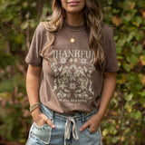 Person outside wearing a brown t-shirt with 'Thankful in All Seasons' design and floral elements.