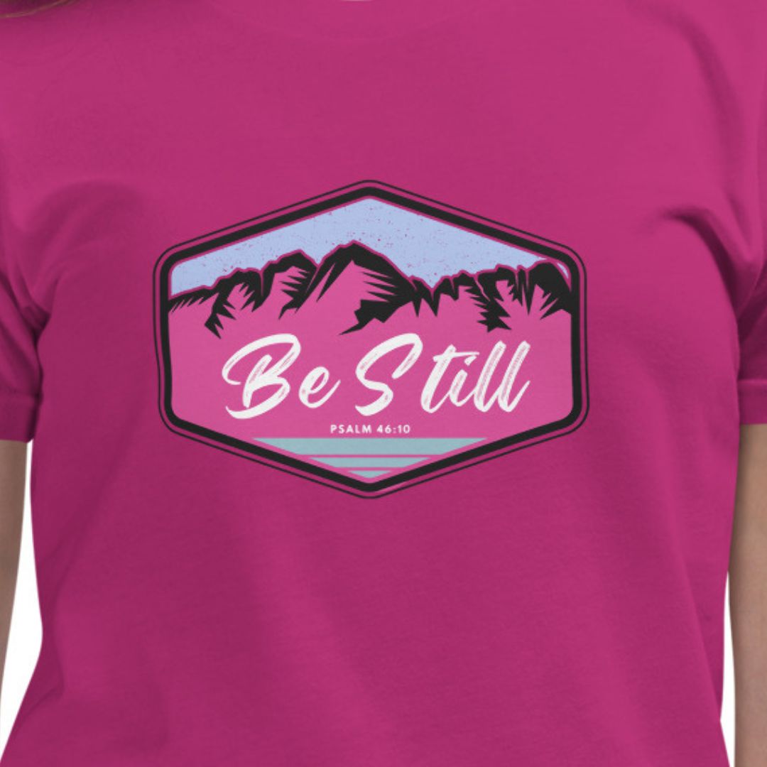 Pink t-shirt with 'Be Still' graphic and mountain design on a white background