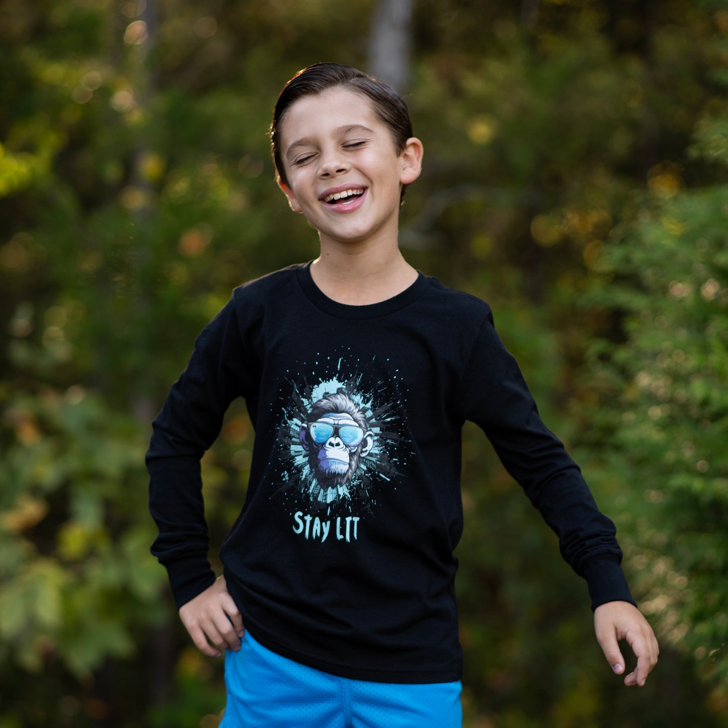 Child wearing a black long-sleeve shirt while laughing with a graphic design and text, standing outdoors with greenery in the background.