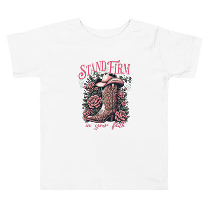 Christian graphic tee featuring a cowboy boot and floral design with the inspirational phrase “Stand Firm in Your Faith” on a pink background. Includes Bible verse 1 Corinthians 16:13 inscribed on the hat. Faith-based western apparel on white t-shirt.