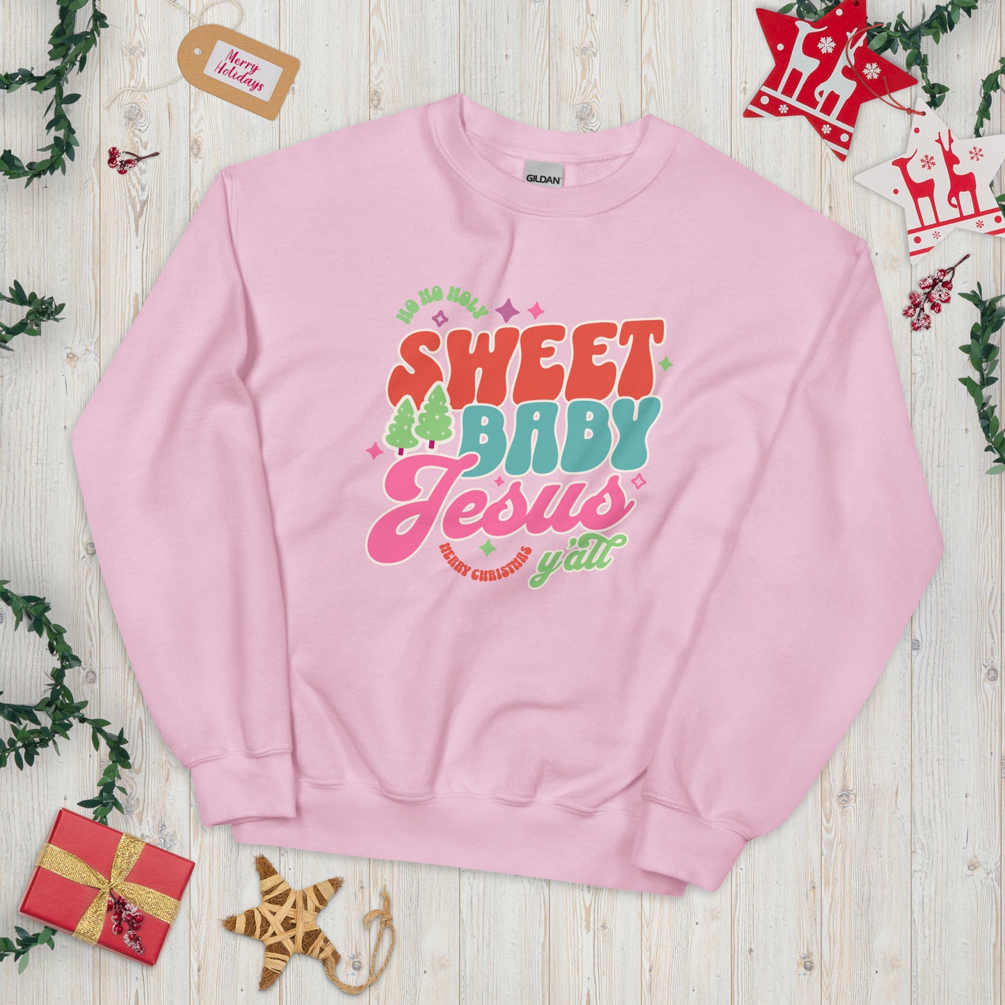 Light pink Christmas sweatshirt displayed flat on a light wood plank background, surrounded by festive decorations including ornaments, greenery, and holiday accents.