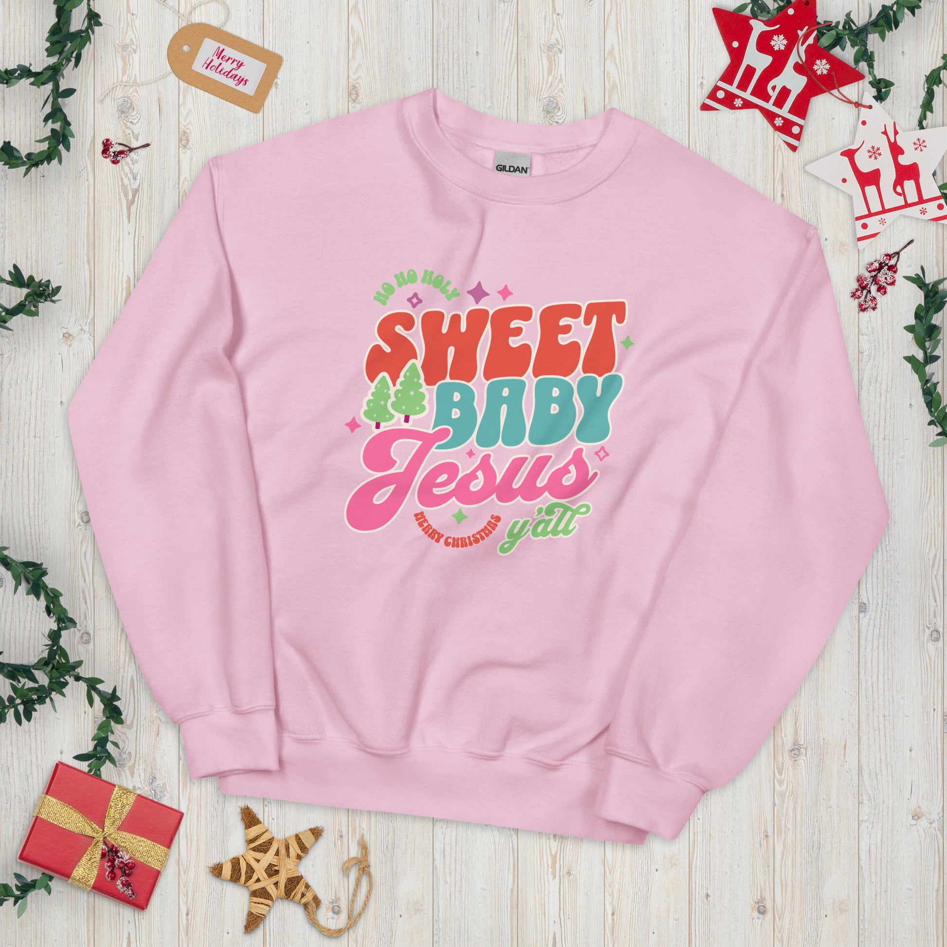 Light pink Christmas sweatshirt displayed flat on a light wood plank background, surrounded by festive decorations including ornaments, greenery, and holiday accents.
